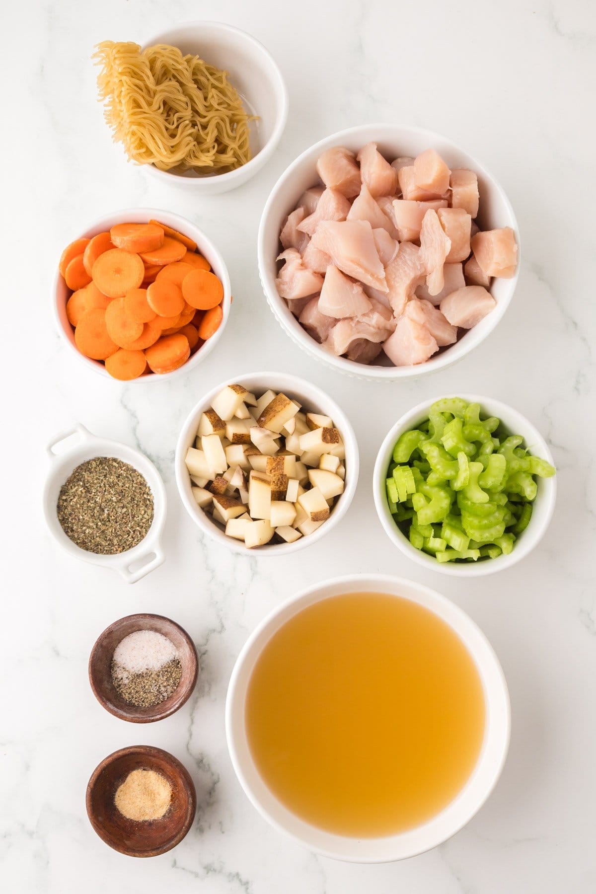 An overhead view of ingredients for slow cooker chicken soup: uncooked noodles, diced raw chicken, sliced carrots, chopped mushrooms, celery, dried herbs, chicken broth, salt, and pepper—all arranged on a white surface.