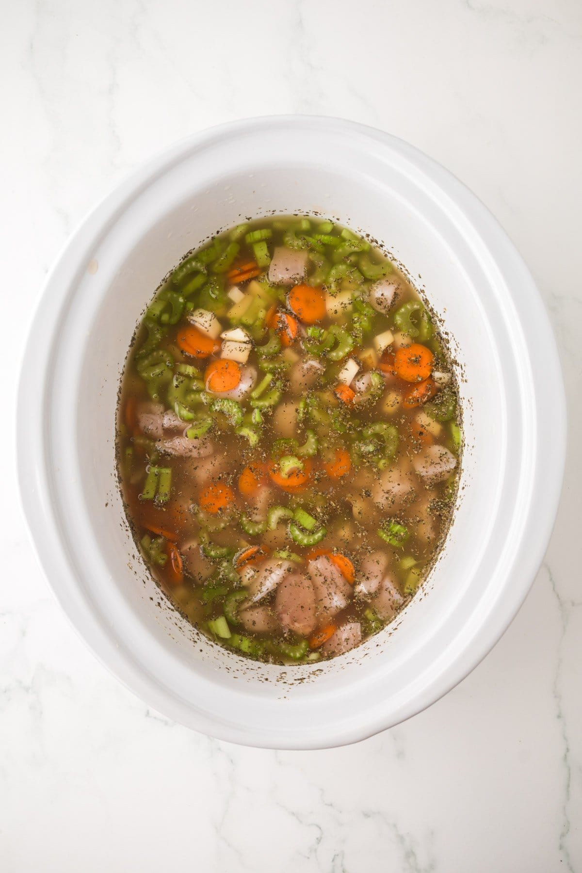 A white slow cooker filled with slow cooker chicken soup—broth, sliced carrots, celery, chopped chicken, and seasonings—sits on a light marble countertop.