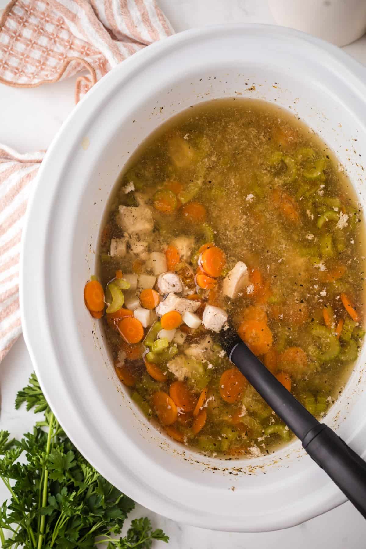 A slow cooker chicken soup filled with carrots, celery, and broth simmers gently. A black ladle rests in the soup. Fresh parsley and a striped towel are nearby on a white countertop.