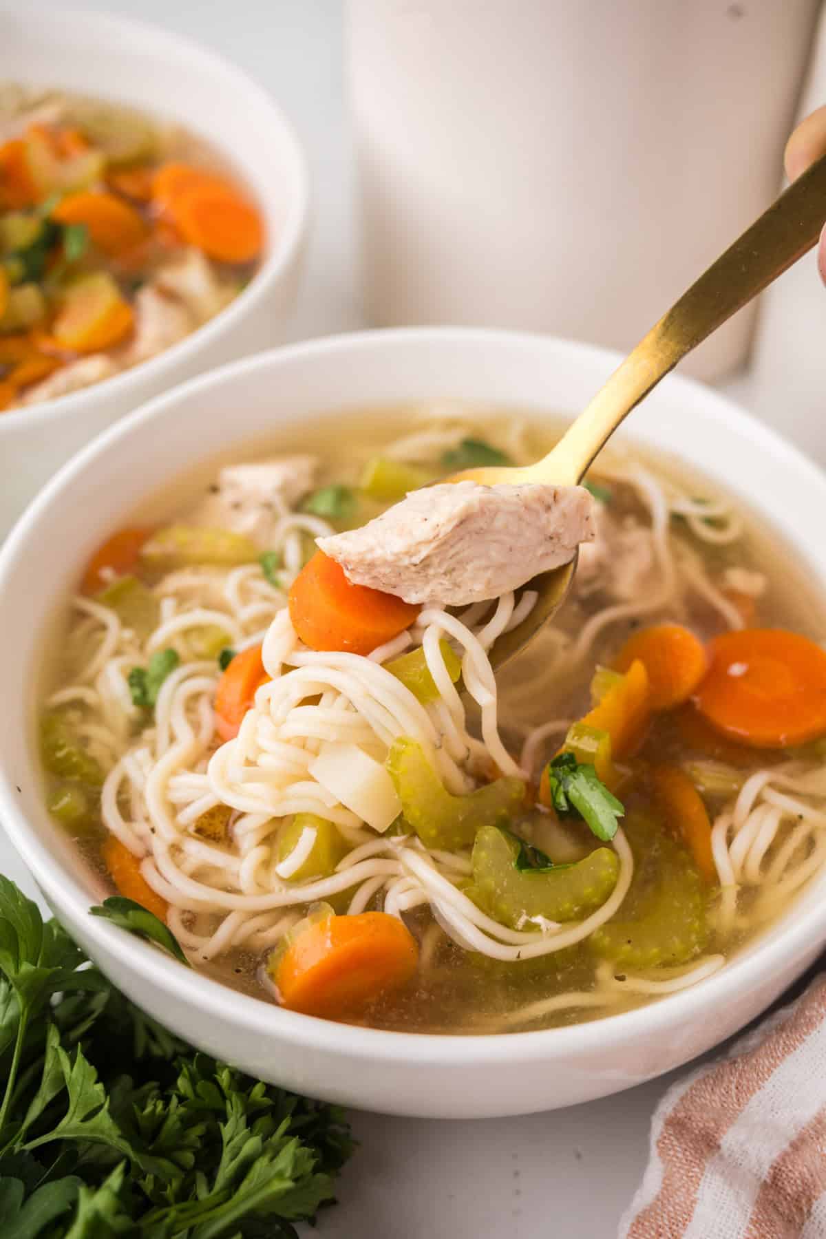 A bowl of slow cooker chicken soup with carrots, celery, and noodles. A gold spoon lifts up a bite with chicken, noodles, and vegetables. Fresh parsley and a striped napkin are nearby.