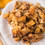 A white plate holds a serving of baked French toast casserole made from golden-brown, chunky bread pieces, with a syrupy glaze and crispy edges. A yellow cloth napkin is partially visible in the upper left corner.