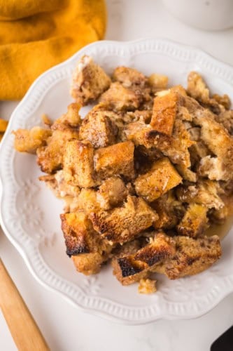 A white plate holds a serving of baked French toast casserole made from golden-brown, chunky bread pieces, with a syrupy glaze and crispy edges. A yellow cloth napkin is partially visible in the upper left corner.