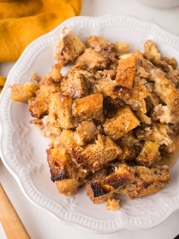 A white plate holds a serving of baked French toast casserole made from golden-brown, chunky bread pieces, with a syrupy glaze and crispy edges. A yellow cloth napkin is partially visible in the upper left corner.
