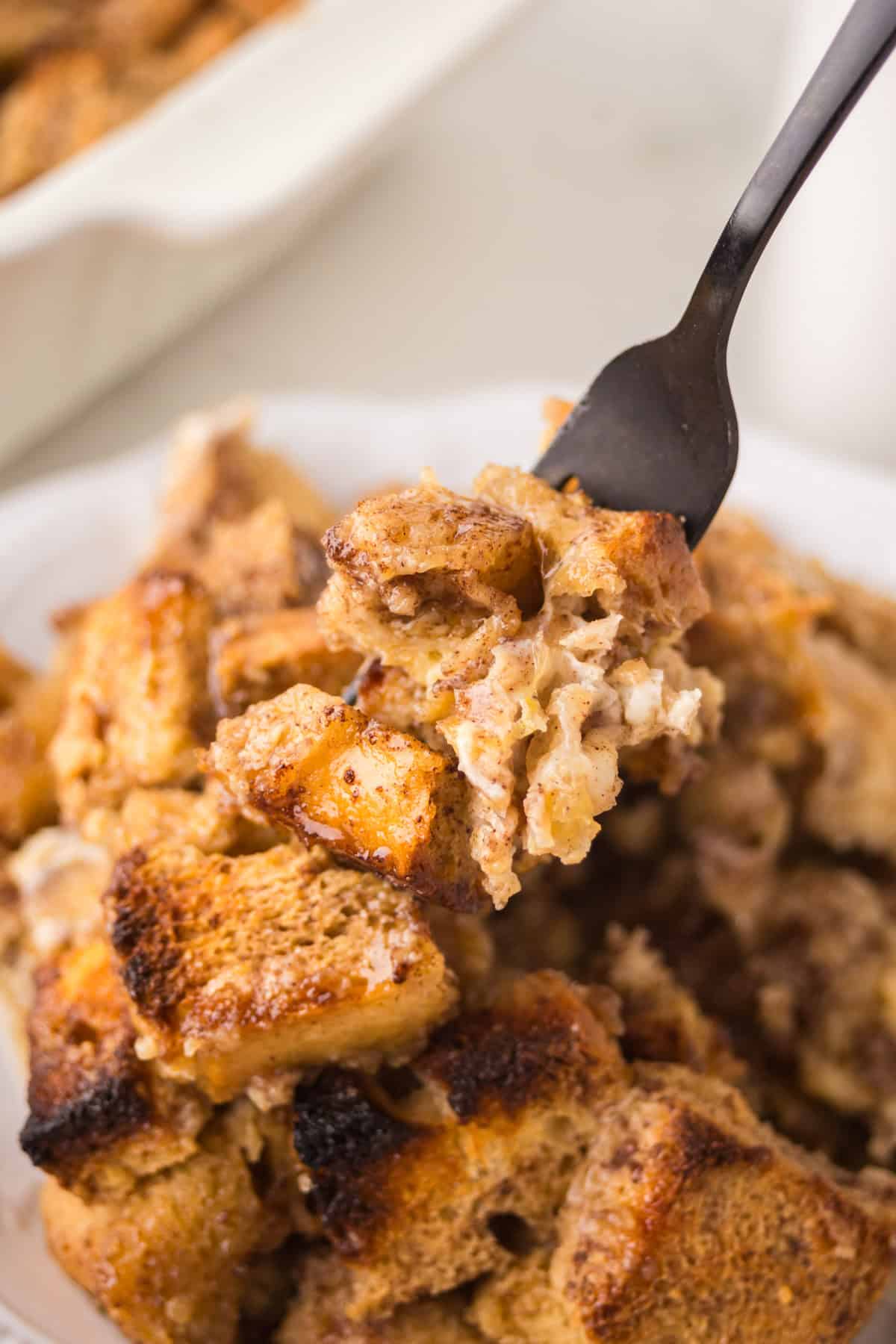 A close-up of a fork holding a bite of bread pudding above a plate filled with more bread pudding pieces, showing a golden brown, soft, and slightly crispy texture.