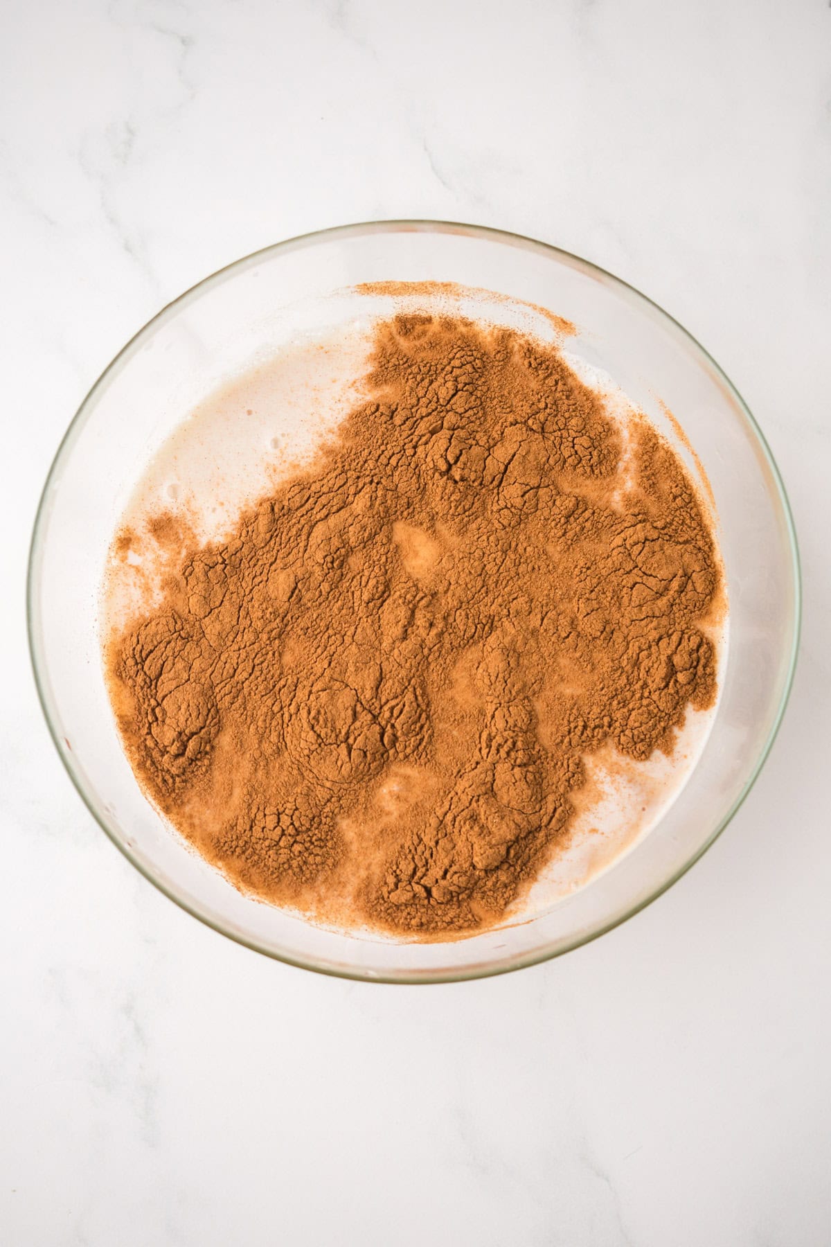 A clear glass bowl filled with a white mixture, likely milk, topped with a generous amount of brown cocoa powder, on a white marble surface.