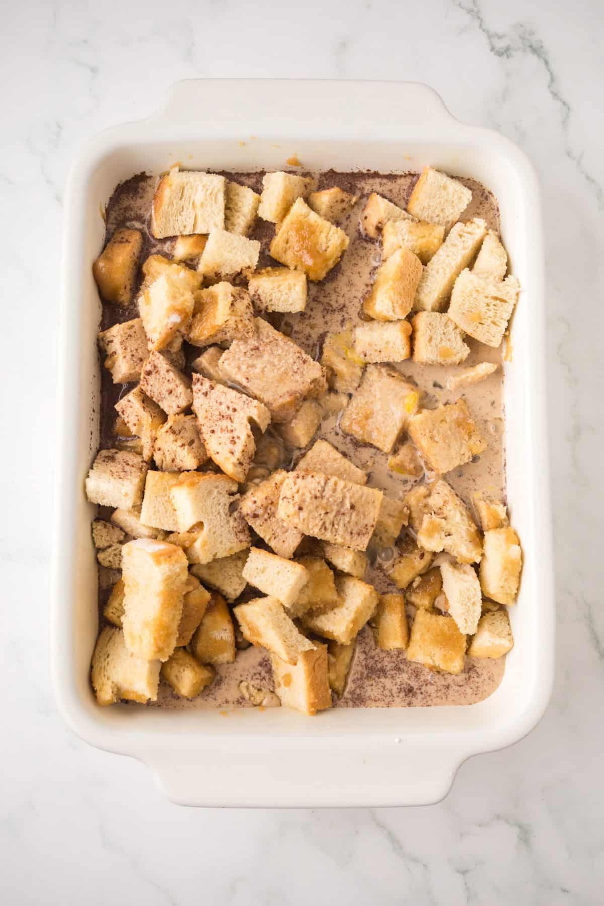 Cubed bread pieces soaked in a creamy, speckled liquid mixture in a white rectangular baking dish, placed on a light marble surface.