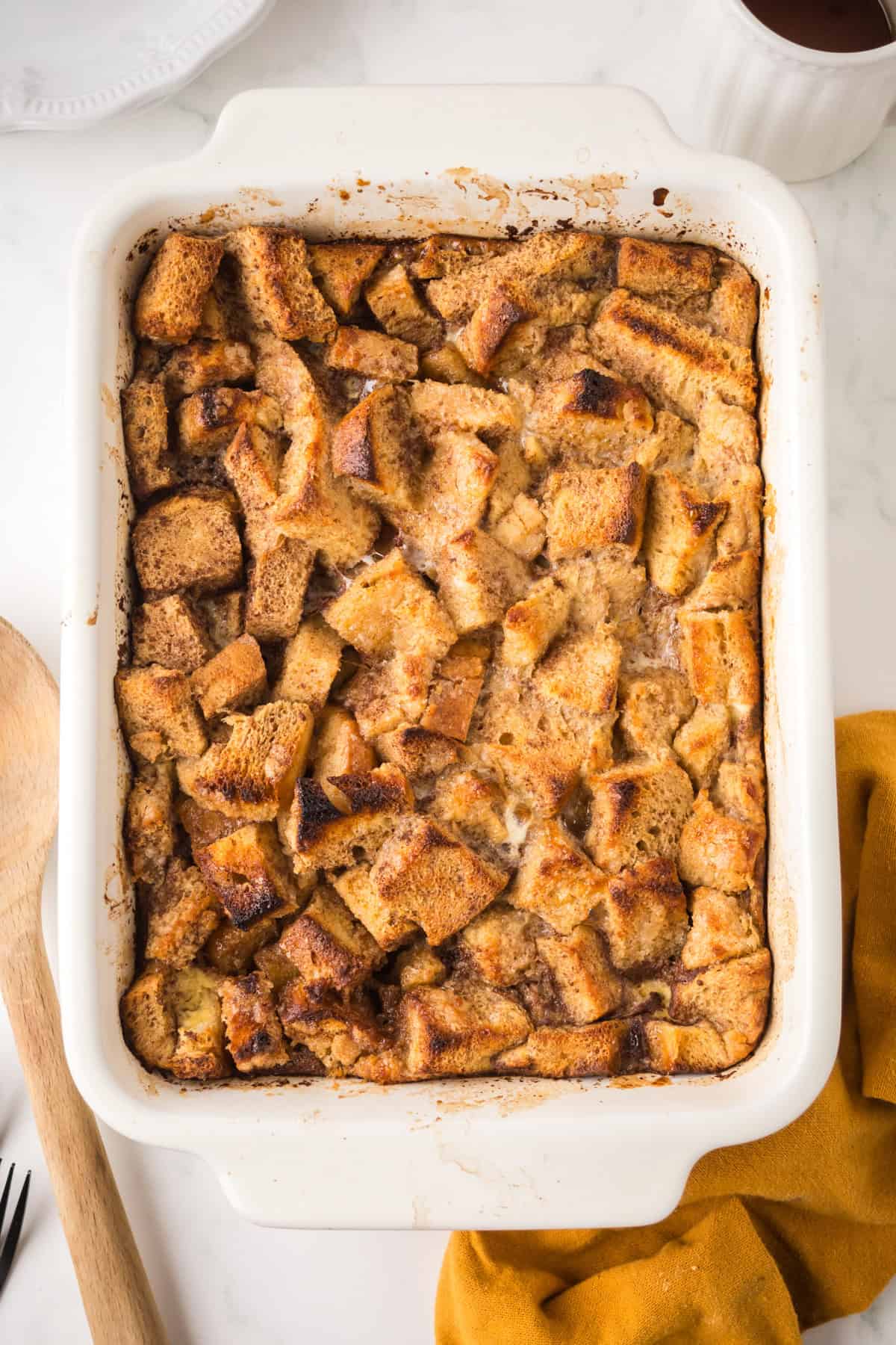 A white baking dish filled with golden-brown bread pudding, featuring toasted, uneven cubes of bread. A wooden spoon, yellow cloth, and part of a white plate are visible nearby.