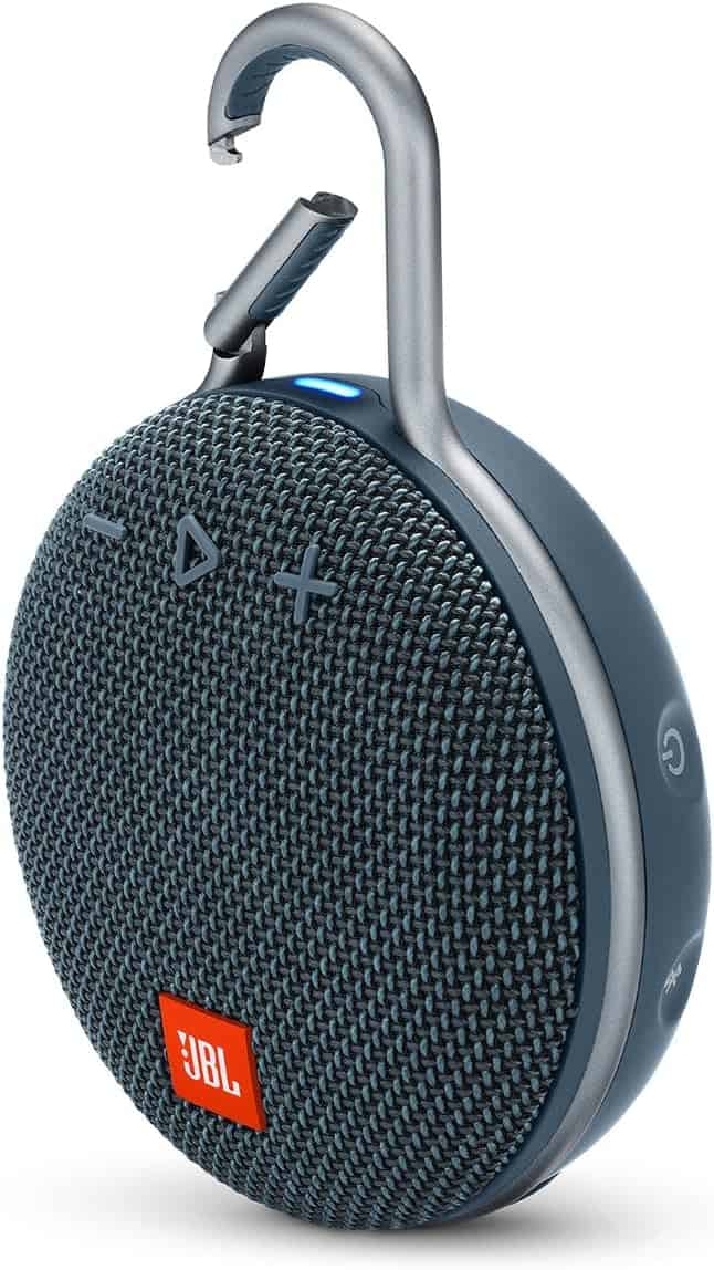 A portable JBL Bluetooth speaker with a fabric front, control buttons, and an integrated metal carabiner for easy attachment. The speaker is mostly dark blue and features the orange JBL logo.