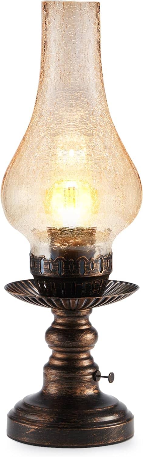 An antique-style oil lamp with a bronze base and a tall, crackled glass chimney, glowing warmly with a lit bulb inside.