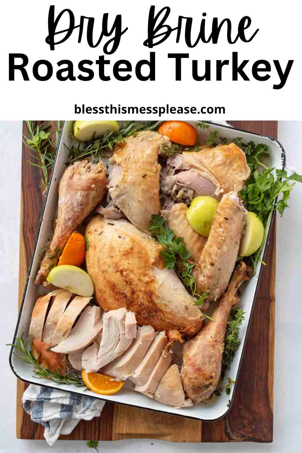 A large dry brine turkey, carved and arranged in a white pan, is garnished with fresh herbs, green apple slices, and orange wedges. The pan rests on a wooden board with a striped kitchen towel beneath.