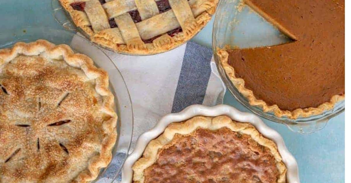 Four different pies on a blue surface—including one with a sugared crust, perfect for learning how to make pie crust—one with a lattice top, one missing a slice of pumpkin pie, and another with a crinkly top in a white dish surround a striped cloth.