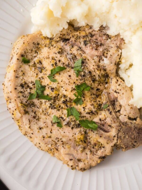A seasoned, cooked lemon pepper pork chop garnished with parsley, served next to a portion of creamy mashed potatoes on a white plate.