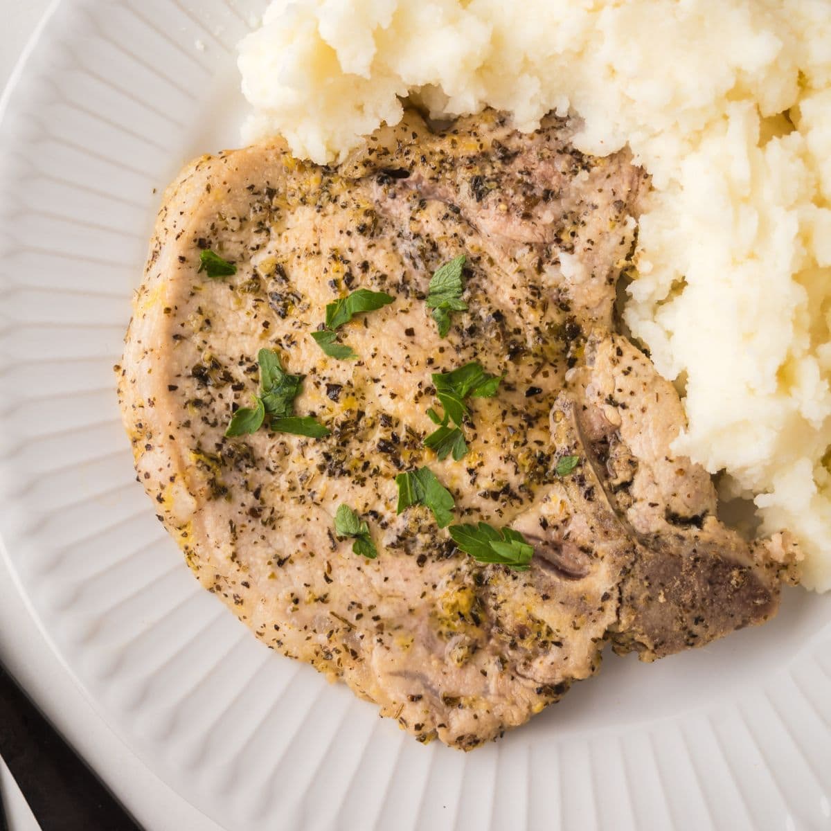 A seasoned, cooked lemon pepper pork chop garnished with parsley, served next to a portion of creamy mashed potatoes on a white plate.