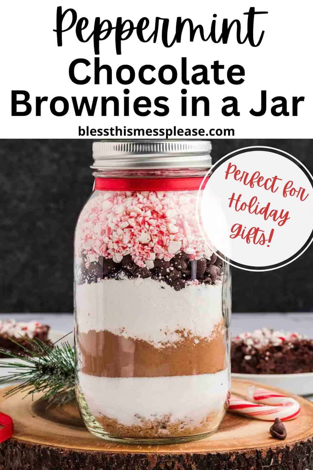 A mason jar filled with layered peppermint chocolate brownies ingredients, including cocoa, chocolate chips, and crushed peppermint, sits on a festive wooden surface with a pine sprig. The label reads Peppermint Chocolate Brownies in a Jar.