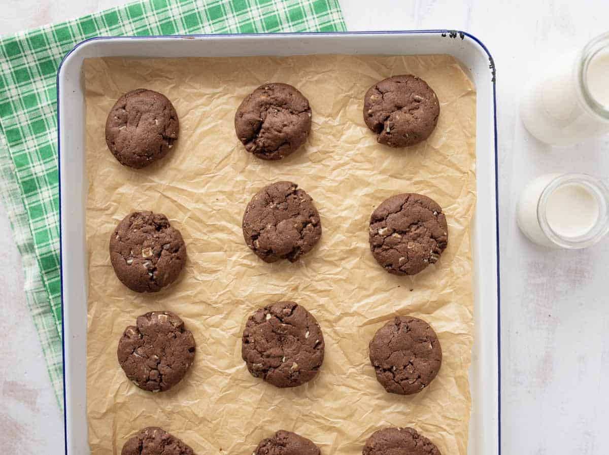 A baking tray lined with parchment paper holds twelve chocolate cookies. Two small bottles of milk and a green checkered cloth are nearby on a white surface.
