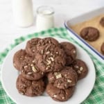 A white plate filled with andes mint chocolate cookies topped with chopped nuts sits on a green checkered cloth, with a baking tray of cookies and two bottles of milk in the background.