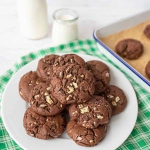 A white plate filled with andes mint chocolate cookies topped with chopped nuts sits on a green checkered cloth, with a baking tray of cookies and two bottles of milk in the background.