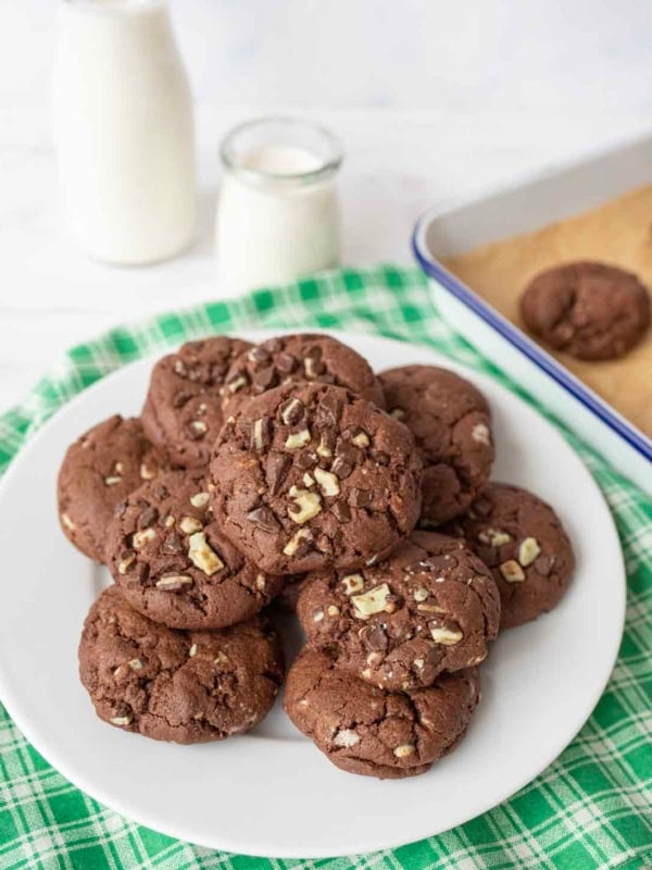 A white plate filled with andes mint chocolate cookies topped with chopped nuts sits on a green checkered cloth, with a baking tray of cookies and two bottles of milk in the background.
