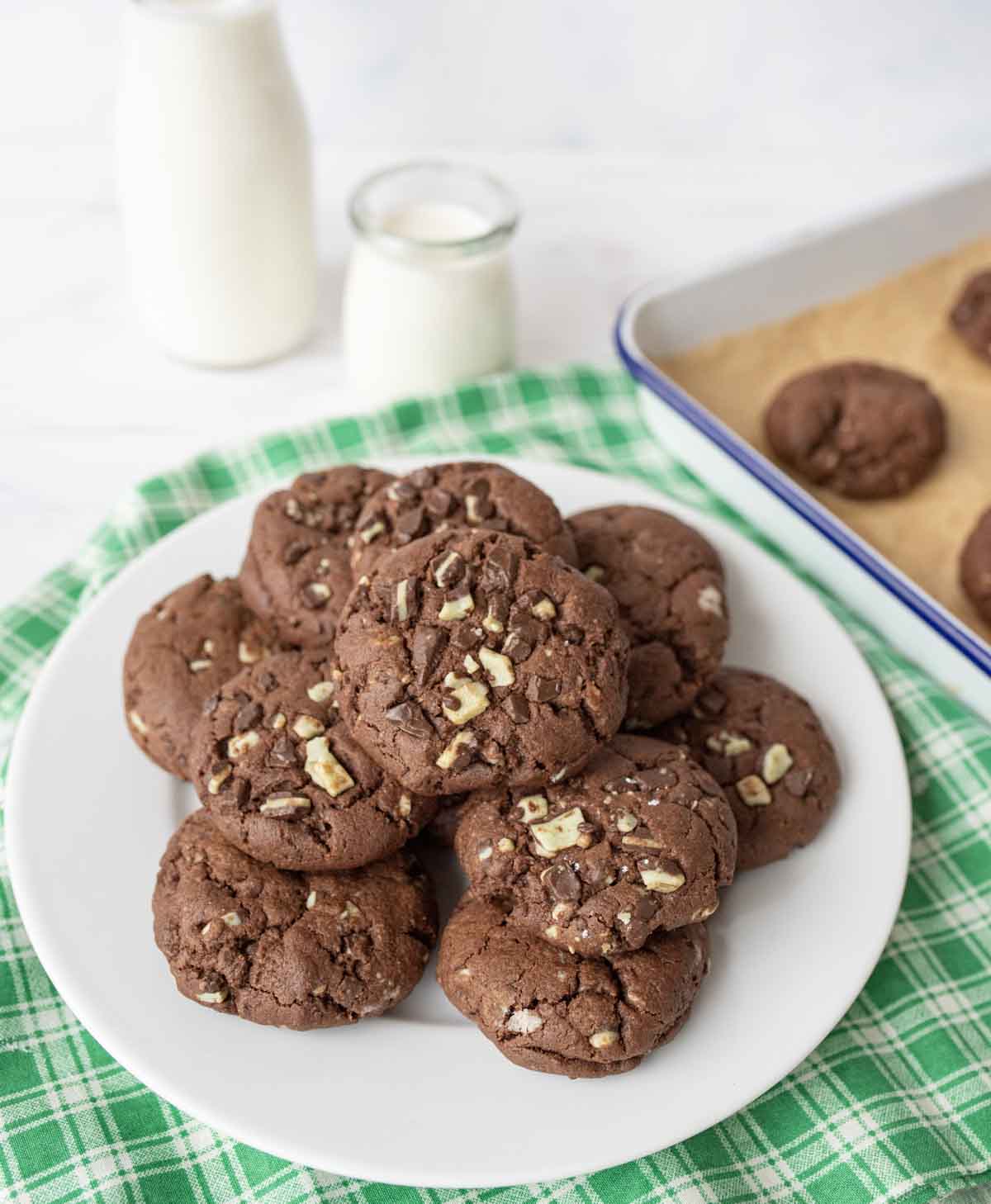 A white plate filled with andes mint chocolate cookies topped with chopped nuts sits on a green checkered cloth, with a baking tray of cookies and two bottles of milk in the background.