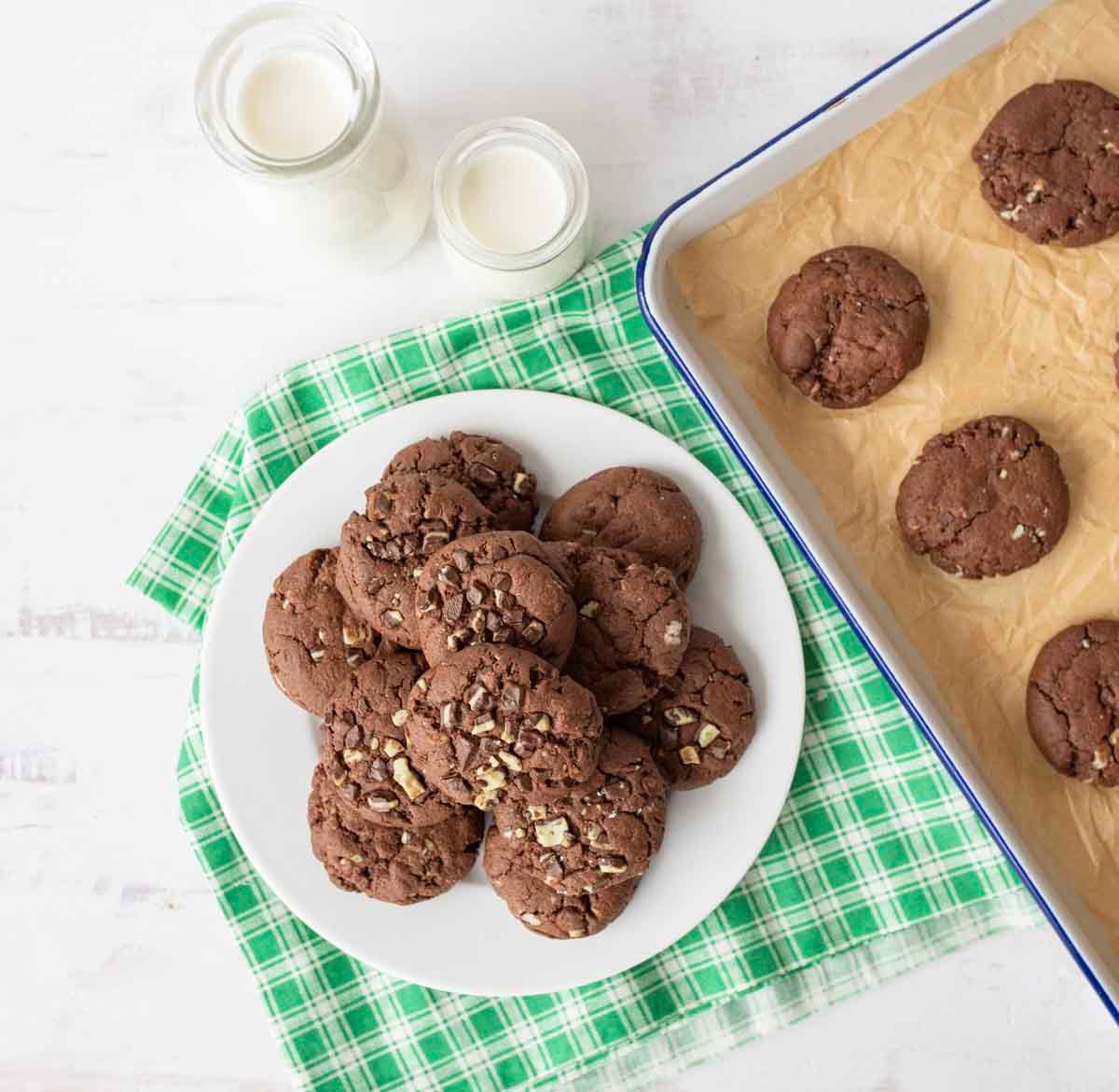 A plate of chocolate chip cookies on a green plaid cloth, next to a baking tray with more cookies and two small bottles of milk on a white surface.