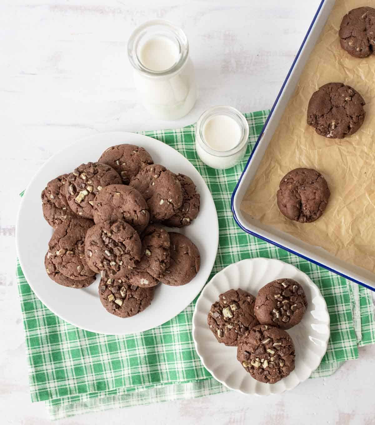 A plate and a small dish of chocolate cookies with chocolate chunks are on a green checkered cloth. Nearby are two glasses of milk and a baking tray lined with parchment paper, holding four more cookies.