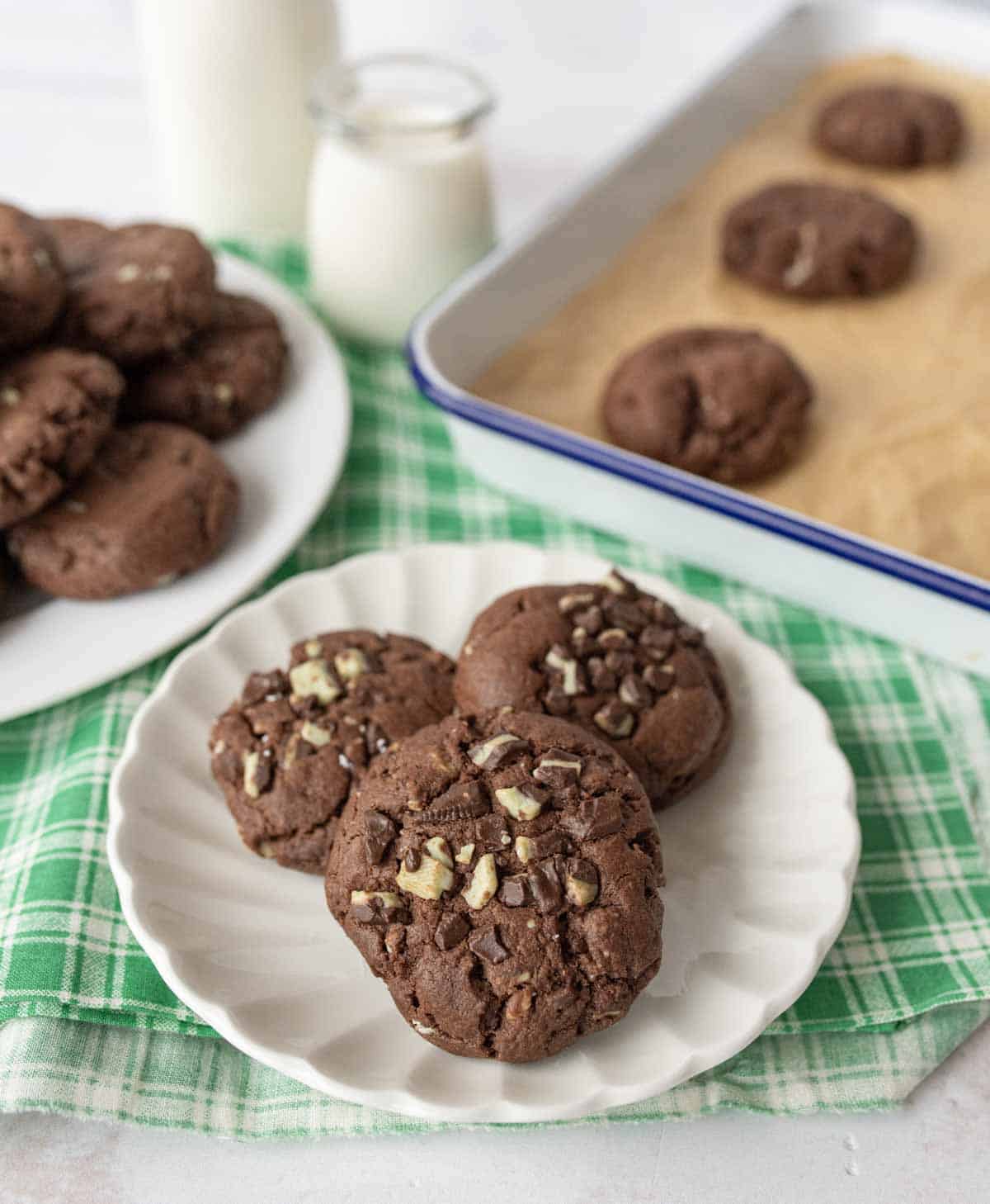 Three chocolate cookies with chocolate chunks are on a white plate. In the background, there are more cookies on a white plate, cookies on a baking tray, and bottles of milk, all on a green checkered cloth.