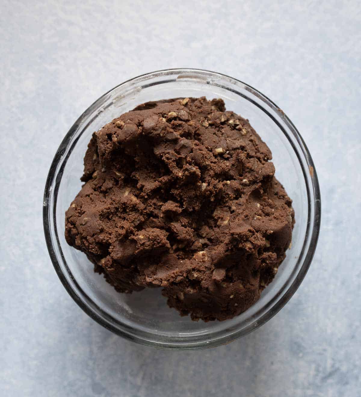A glass bowl filled with a mound of chocolate cookie dough sits on a light gray surface.