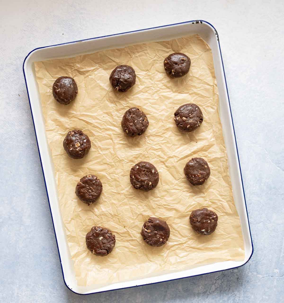 A baking tray lined with crumpled parchment paper holds twelve evenly spaced balls of chocolate cookie dough, ready to be baked. The background is a light, textured surface.