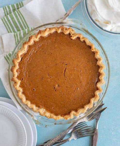 A whole baked pumpkin pie in a glass dish sits on a blue surface with a green-striped towel, a bowl of whipped cream, white plates, silver utensils, and tips on how to make pie crust nearby.