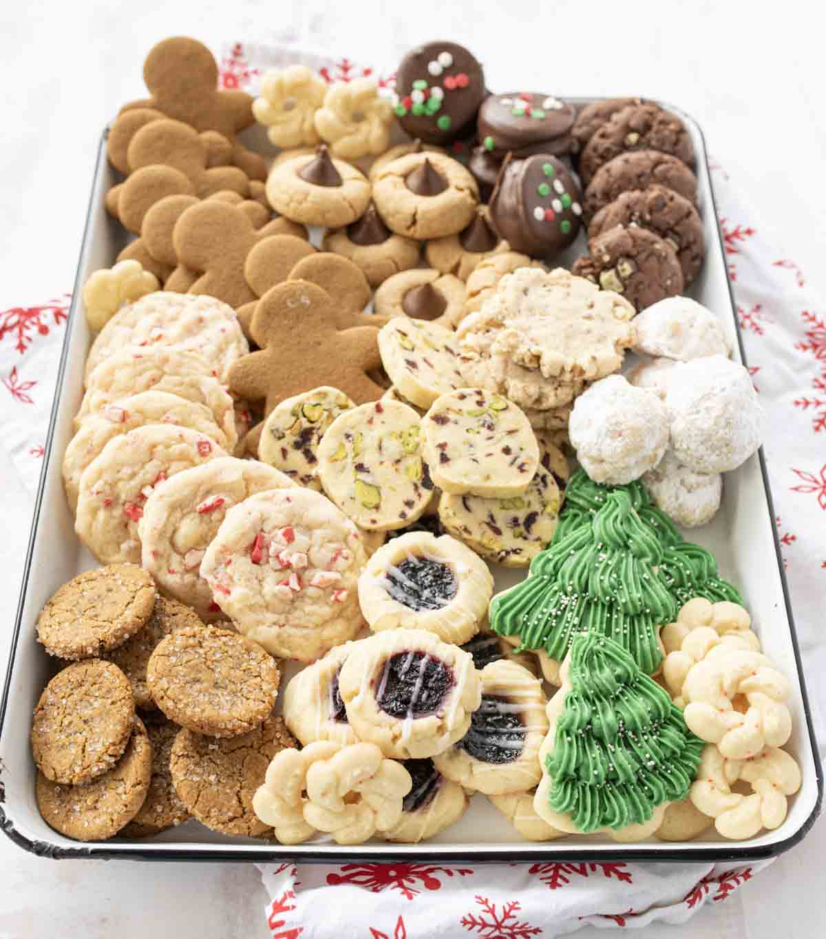 A large tray filled with a variety of holiday cookies, including gingerbread men, chocolate cookies with sprinkles, powdered sugar snowballs, jam-filled cookies, green Christmas tree cookies, and more.