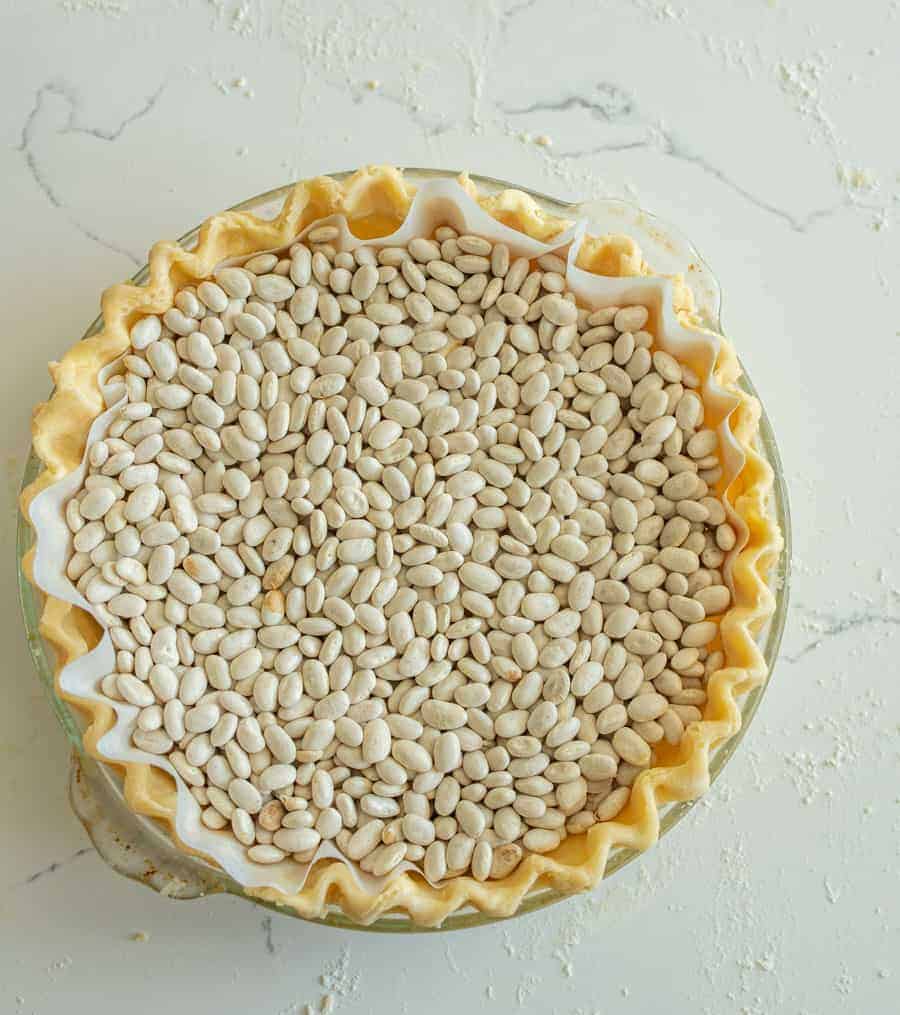 Unbaked pie crust in a dish lined with parchment paper and filled with dried white beans for blind baking, demonstrating how to make pie crust on a light-colored surface.