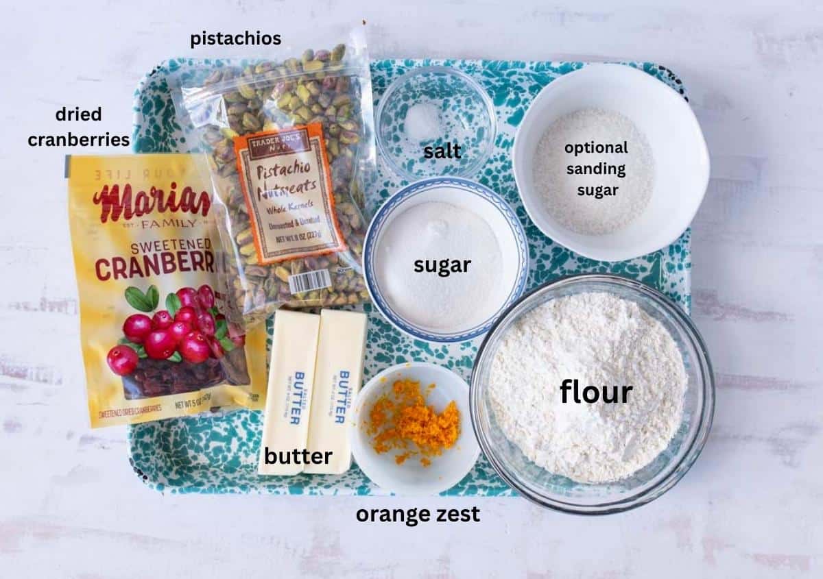 A tray displays labeled ingredients for baking: dried cranberries, pistachios, salt, sugar, optional sanding sugar, flour, two sticks of butter, and orange zest in small bowls.