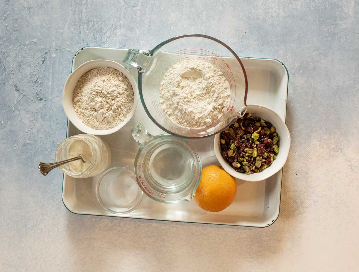 A white tray holds bowls of flour, chopped nuts, and a jar with a spoon, along with a measuring cup of water, a small glass cup, and a whole orange on a light surface.