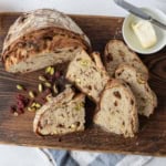 A sliced loaf of bread with visible nuts and dried fruit sits on a wooden cutting board, next to a knife, an orange, scattered pistachios and cranberries, and a small dish of butter with a knife.
