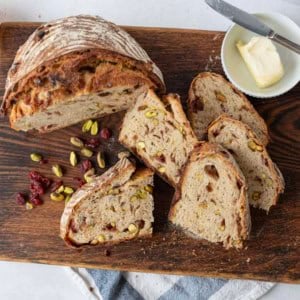 A sliced loaf of bread with visible nuts and dried fruit sits on a wooden cutting board, next to a knife, an orange, scattered pistachios and cranberries, and a small dish of butter with a knife.