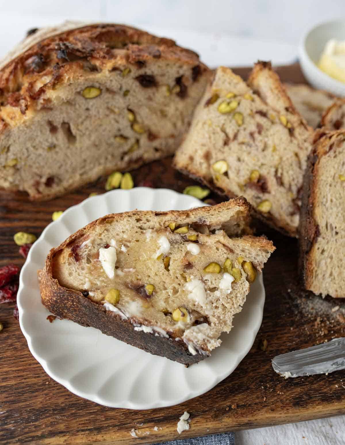 A slice of rustic bread with cranberries and pistachios, spread with butter, rests on a white plate. Behind it, more slices and a loaf sit on a wooden board. A butter knife lies nearby.