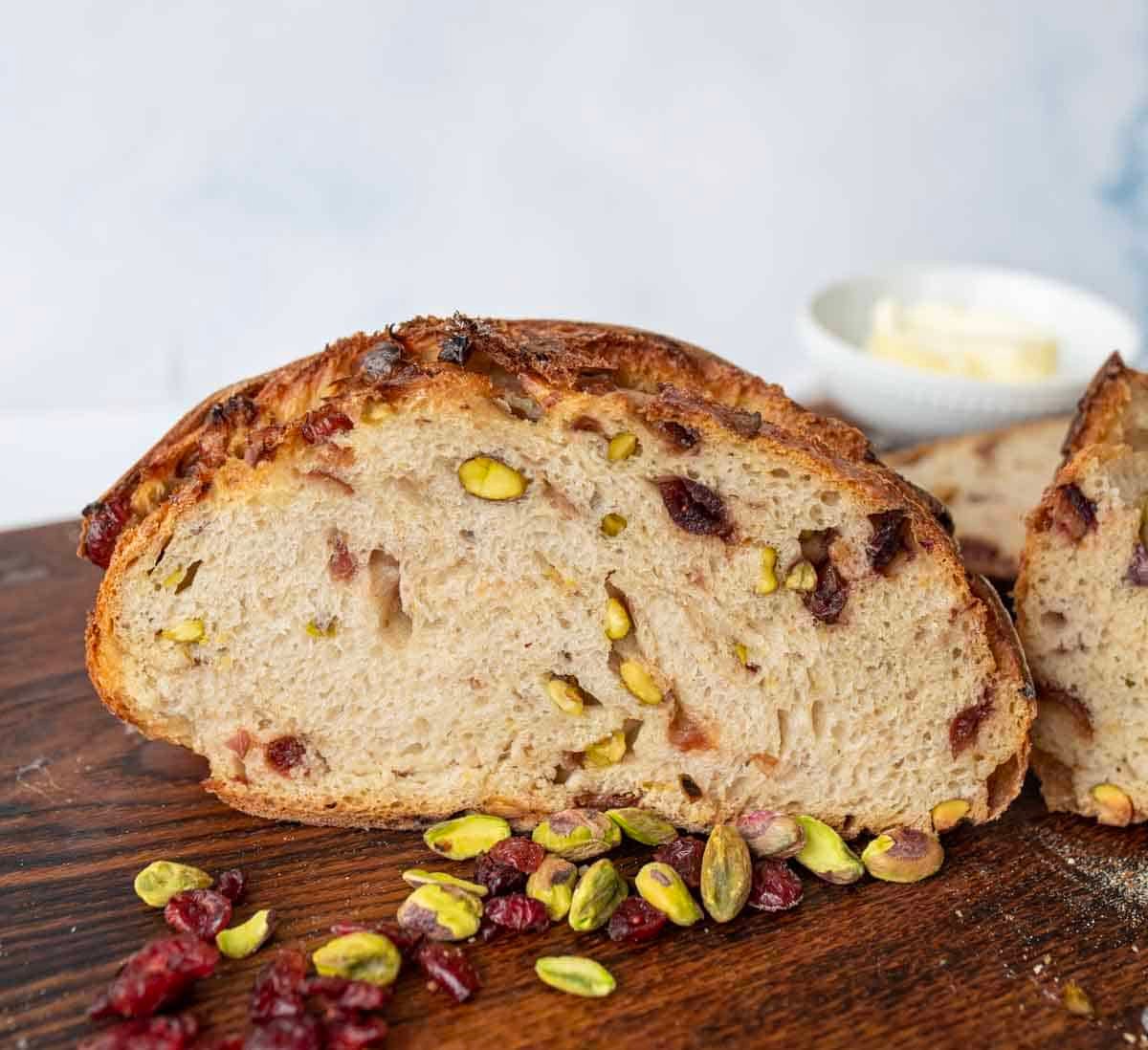 A sliced loaf of rustic bread with visible pistachios and cranberries sits on a wooden board. Scattered pistachios and cranberries are in the foreground; a small bowl of butter is in the background.
