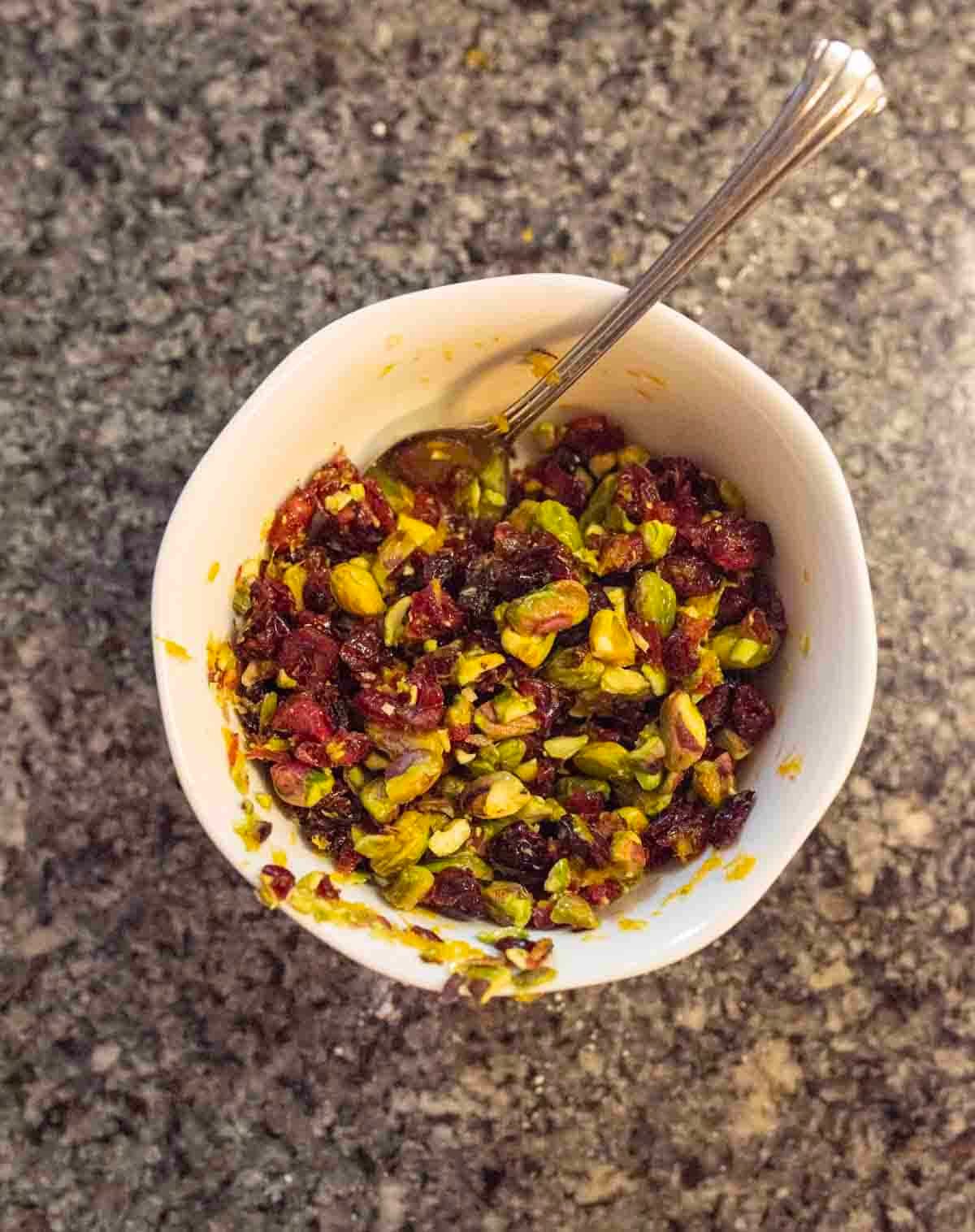 A white bowl filled with a colorful mixture of chopped pistachios, dried cranberries, and orange zest sits on a speckled gray countertop, with a silver spoon resting inside.