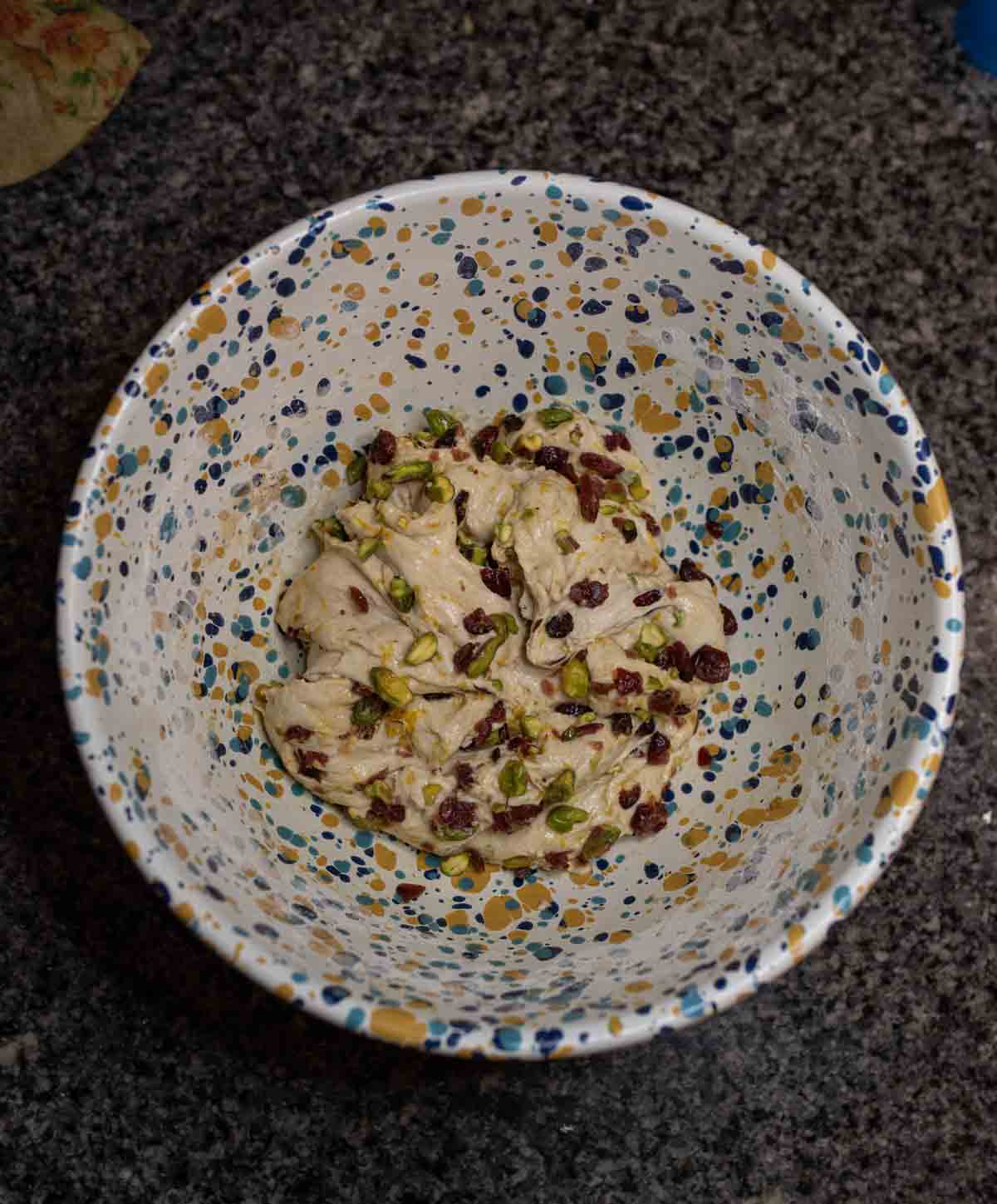 A colorful, speckled bowl contains a mixture topped with chopped pistachios and dried cranberries, resting on a dark countertop.