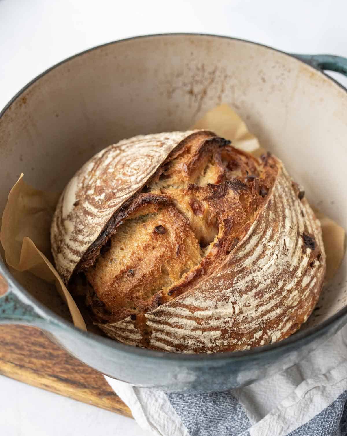 A round loaf of artisanal sourdough bread with a golden, crusty top sits in a Dutch oven lined with parchment paper. The bread has a rustic, swirled pattern and is resting on a wooden board.