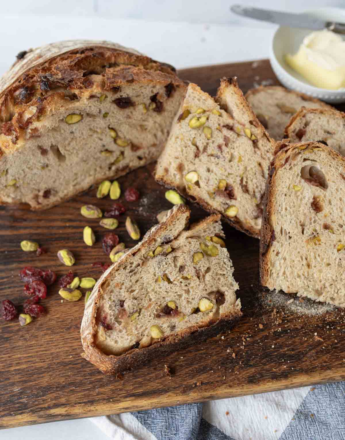 A sliced loaf of rustic cranberry sourdough bread with visible pistachios and cranberries sits on a wooden board, with scattered nuts and berries nearby and a dish of butter in the background.