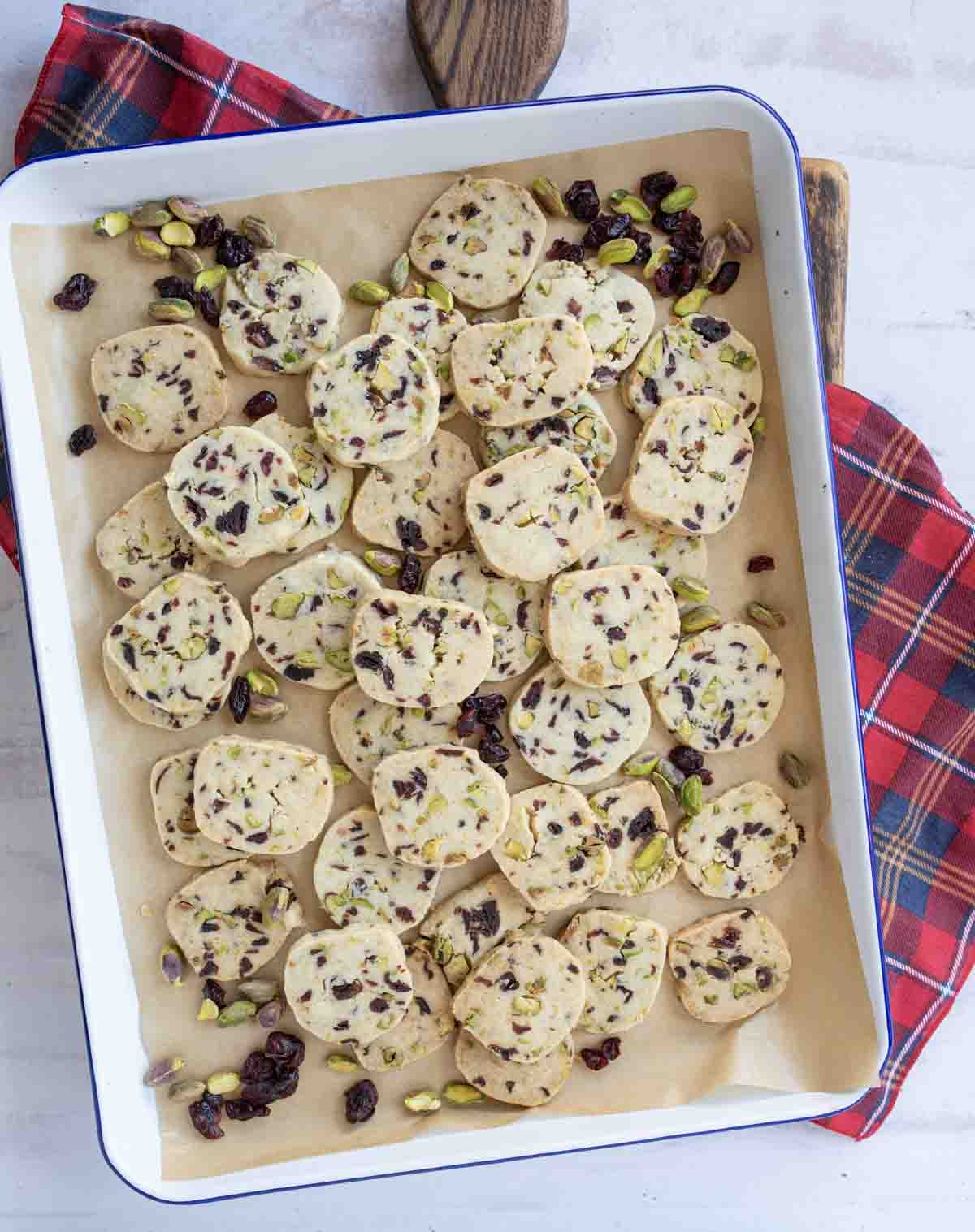 Rectangular white baking tray lined with parchment paper, filled with slices of raw cookie dough studded with pistachios and dried cranberries. Red plaid cloth is partly visible beside the tray.