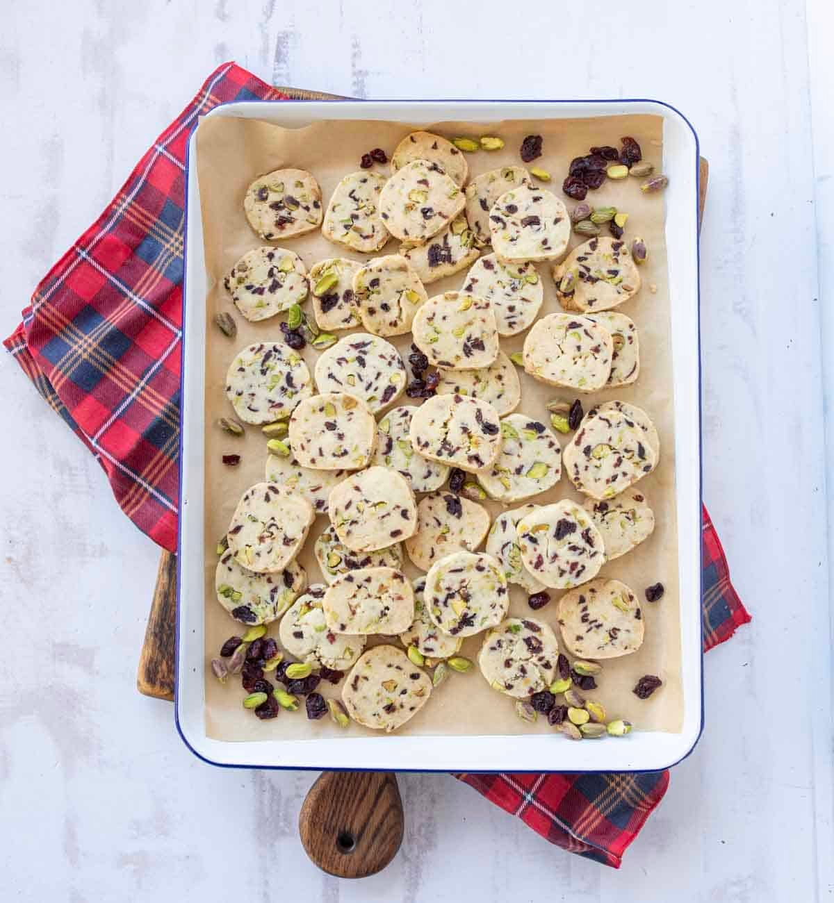 A baking tray lined with parchment paper holds sliced cookies sprinkled with pistachios and dried cranberries, resting on a red plaid cloth atop a light-colored surface.