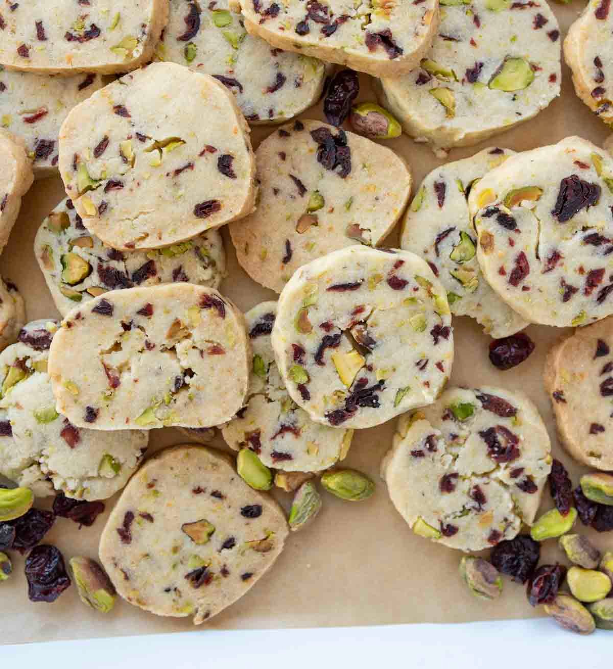 A pile of square and round shortbread cookies with visible pieces of pistachios and dried cranberries on a parchment-lined surface.