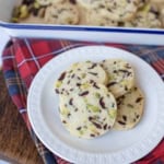 A white plate holds several cranberry orange shortbread cookies, with visible pieces of pistachios and cranberries. More cookies rest in a tray in the background, set on a red plaid cloth over a wooden surface.