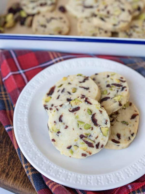 A white plate holds several cranberry orange shortbread cookies, with visible pieces of pistachios and cranberries. More cookies rest in a tray in the background, set on a red plaid cloth over a wooden surface.