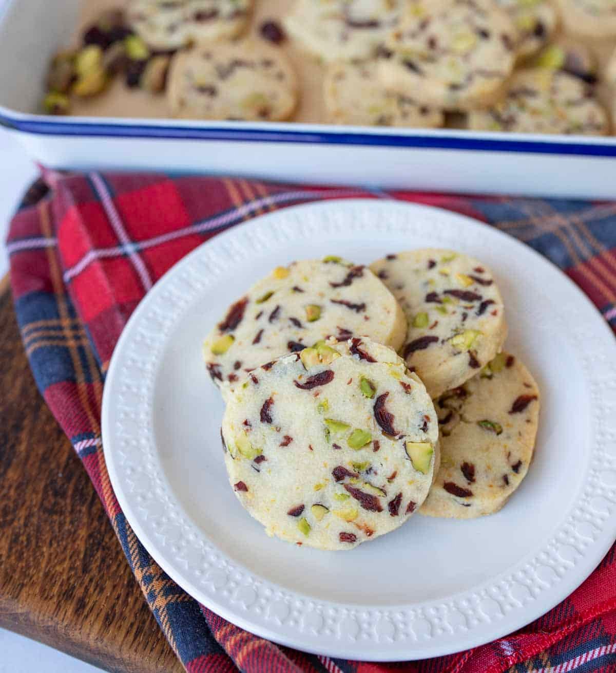 A white plate holds several cranberry orange shortbread cookies, with visible pieces of pistachios and cranberries. More cookies rest in a tray in the background, set on a red plaid cloth over a wooden surface.
