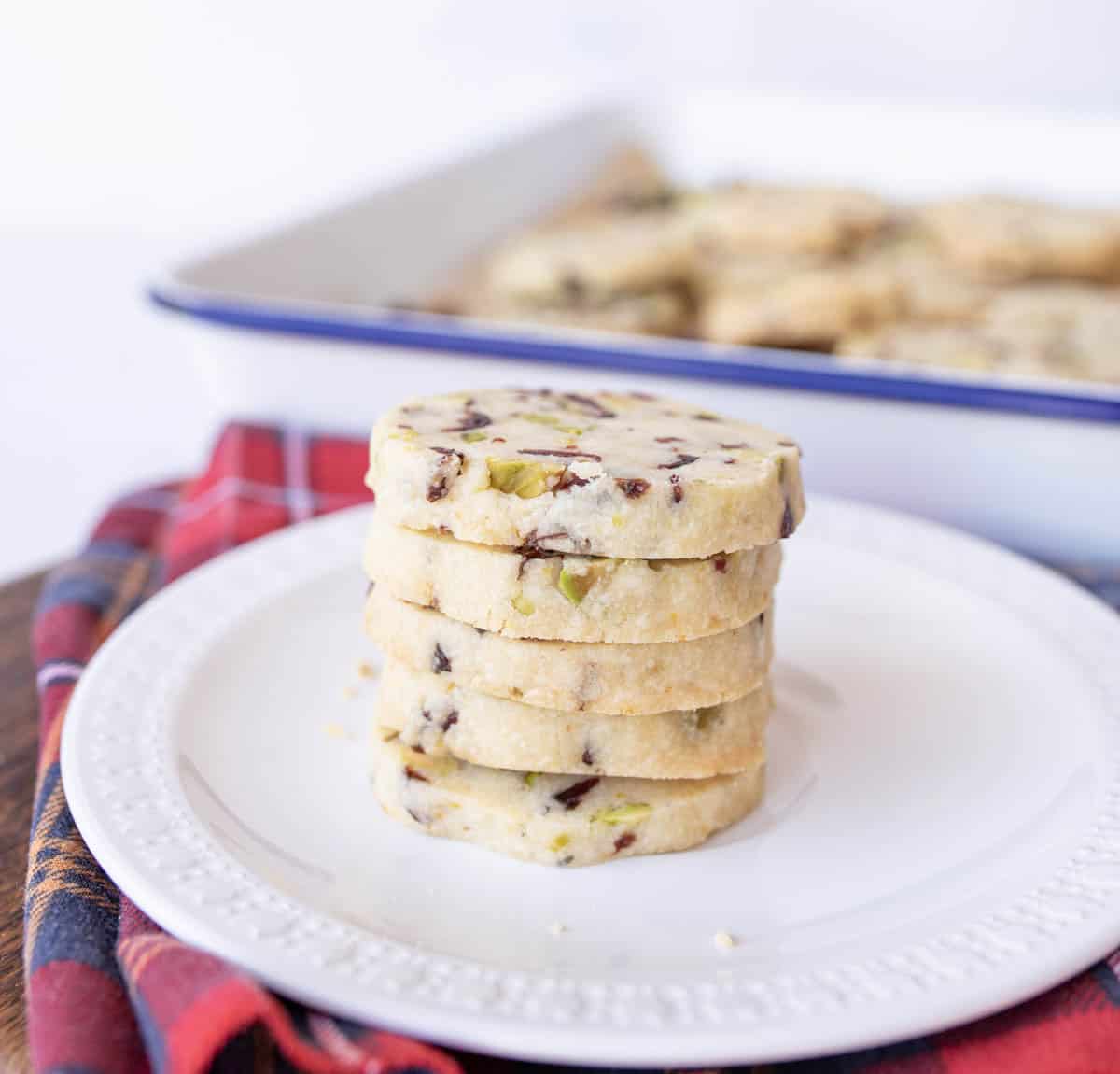 A stack of five round shortbread cookies with bits of pistachio and chocolate sits on a white plate. In the background, more cookies are visible in a rectangular baking dish. A red plaid cloth is beneath the plate.