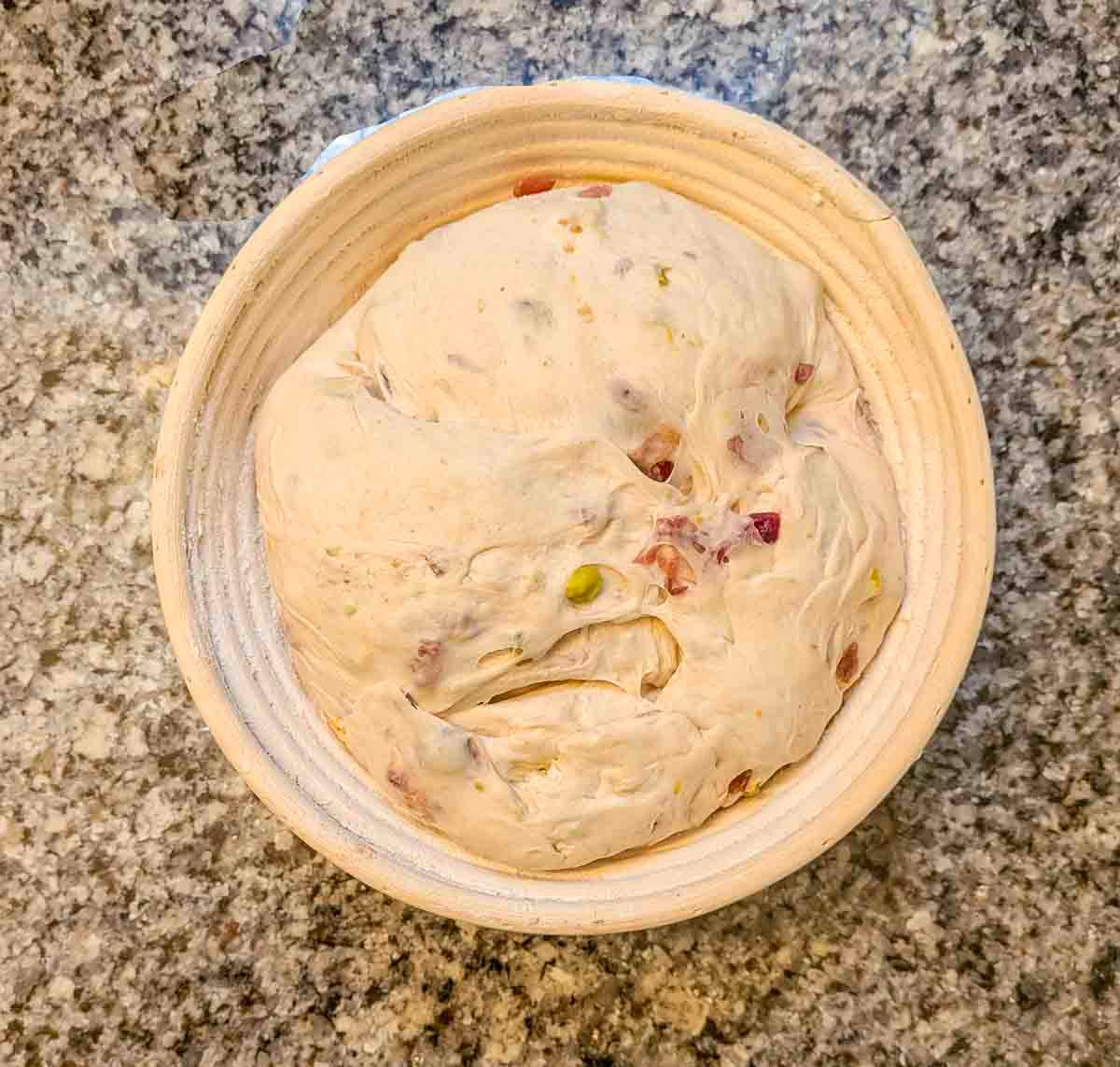 A round bowl filled with risen bread dough containing visible pieces of nuts and dried fruit, placed on a granite countertop.