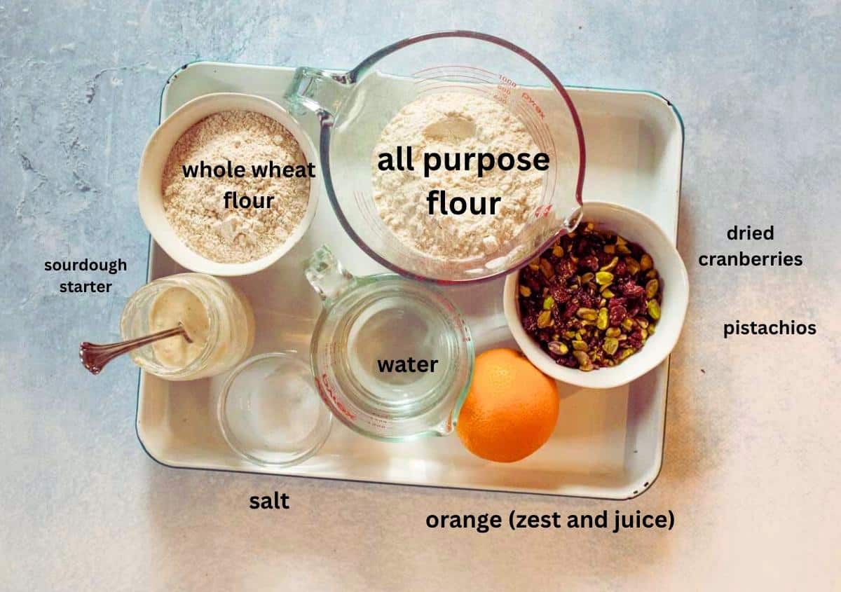 A tray displays bowls of all purpose flour, whole wheat flour, dried cranberries, pistachios, a jar of sourdough starter, a measuring cup of water, salt, and a whole orange for zest and juice.